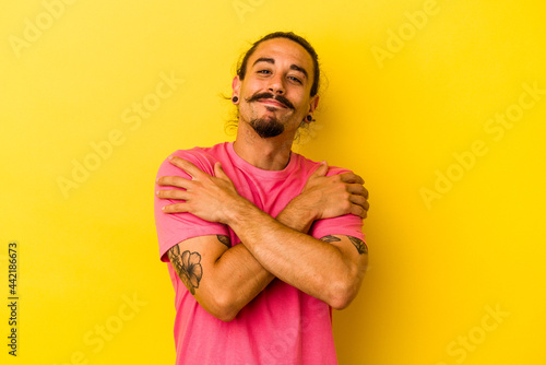 Young caucasian man with long hair isolated on yellow background hugs, smiling carefree and happy. © Asier