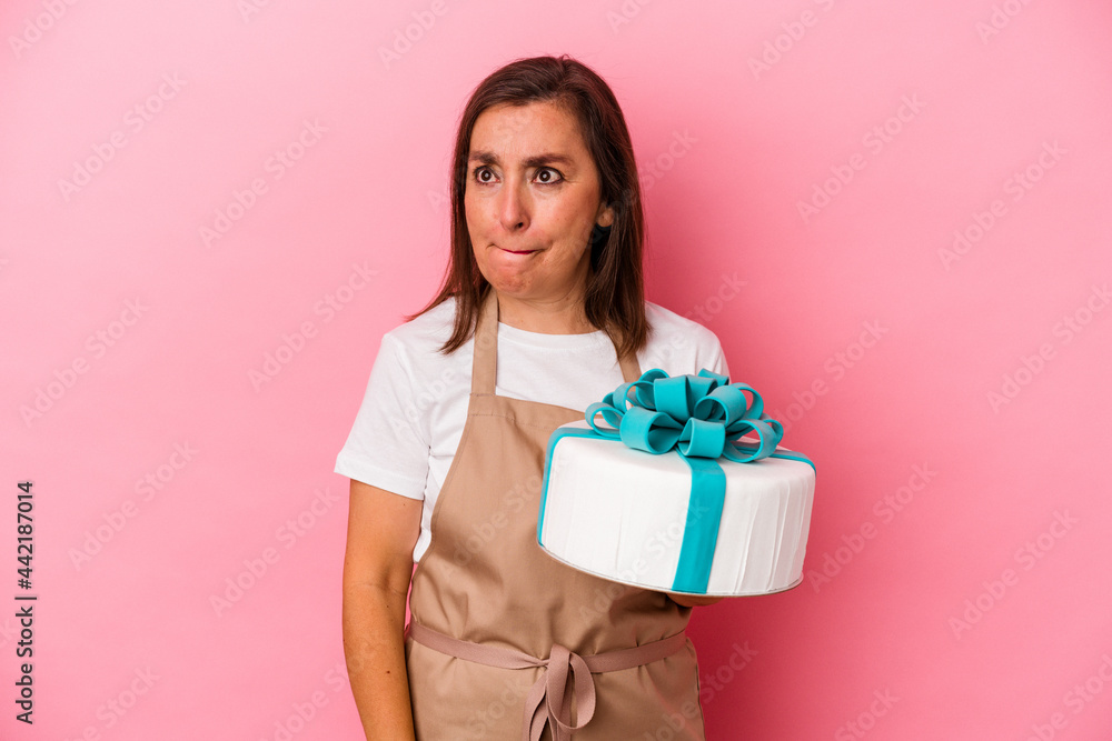 Middle aged pastry chef woman holding a cake isolated on blue background confused, feels doubtful and unsure. Middle aged pastry chef woman holding a cake isolated on blue background confused, feels doubtful and unsure.