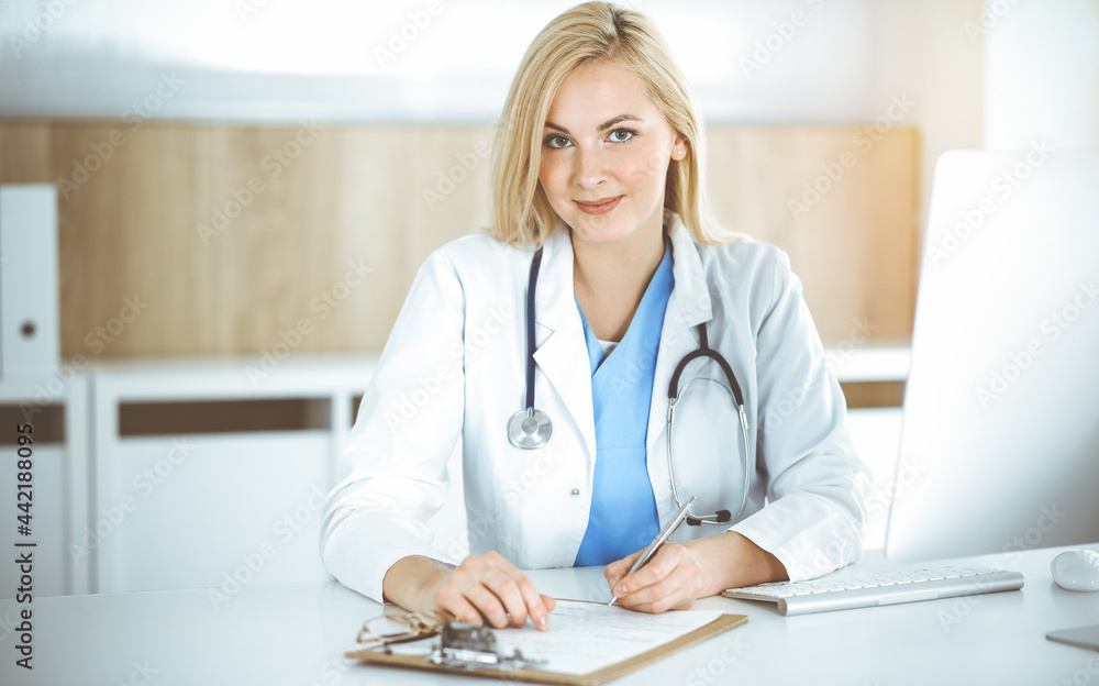 Woman-doctor at work while sitting at the desk in sunny clinic. Blonde cheerful physician filling up medication history record form Woman-doctor at work while sitting at the desk in sunny clinic. Blonde cheerful physician filling up medication history record form