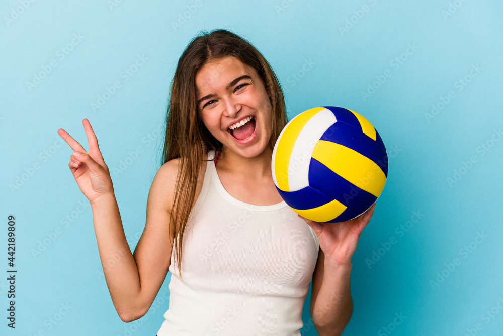 Young caucasian woman playing volleyball isolated on blue background joyful and carefree showing a peace symbol with fingers.