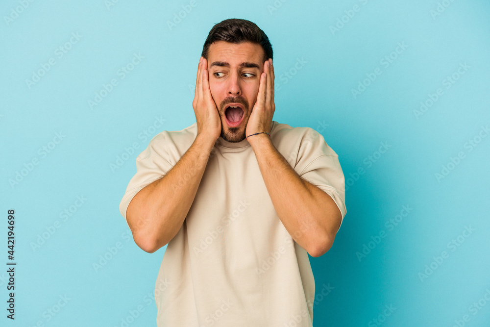 Young caucasian man isolated on blue background covering ears with hands trying not to hear too loud sound. Young caucasian man isolated on blue background covering ears with hands trying not to hear too loud sound.