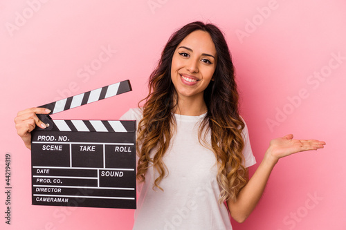 Young mexican woman holding clapperboard isolated on pink background showing a copy space on a palm and holding another hand on waist.