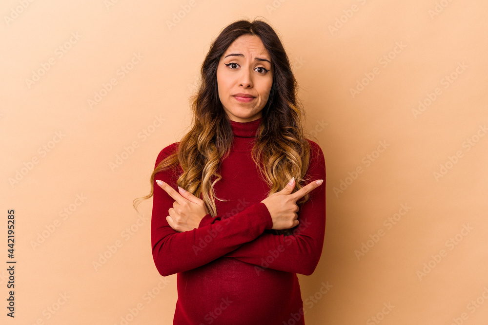 Young mexican pregnant woman isolated on beige background points sideways, is trying to choose between two options. Young mexican pregnant woman isolated on beige background points sideways, is trying to choose between two options.