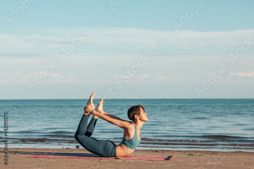 Fototapeta Naklejka Na Ścianę i Meble -  a young attractive woman, of Asian appearance, practicing yoga, performs a stretching exercise, on the beach. the Dhanurasana, the bow pose