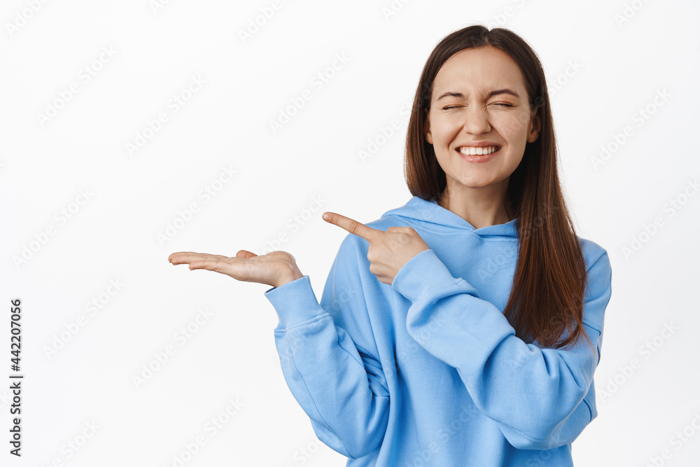 Happy cute girl in hoodie, laughing and smiling, pointing finger at empty open palm, showing something in her hand, standing against white background Happy cute girl in hoodie, laughing and smiling, pointing finger at empty open palm, showing something in her hand, standing against white background