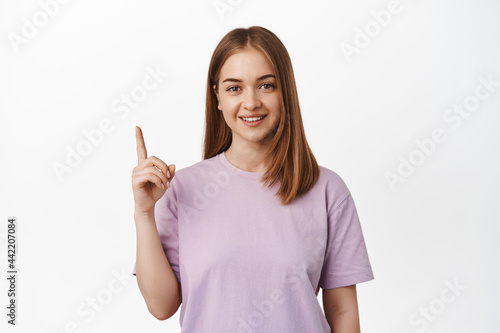 That way. Young smiling woman with natural blond hair, pointing finger up and looking happy, introduce, make announcement, showing banner or logo, white background