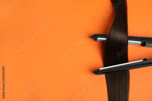 Straightener with brown hair lock on orange background, flat lay. Space for text