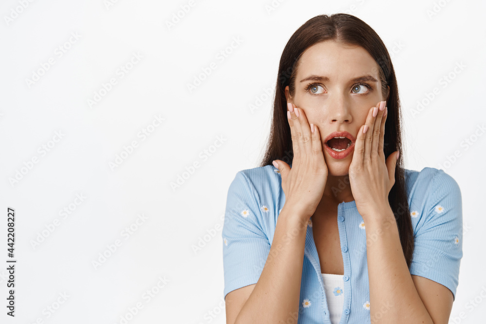 Image of shocked brunette woman gasp, open mouth startled, holding hands on face, staring at upper left corner sale or banner, standing speechless in stupor against white background Image of shocked brunette woman gasp, open mouth startled, holding hands on face, staring at upper left corner sale or banner, standing speechless in stupor against white background