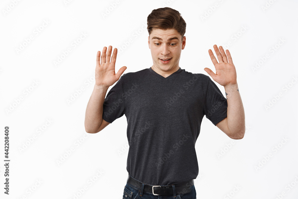 Fototapeta premium Surprised young man drop something, raising hands up and looks down startled, staring below, standing against white background in casual clothes