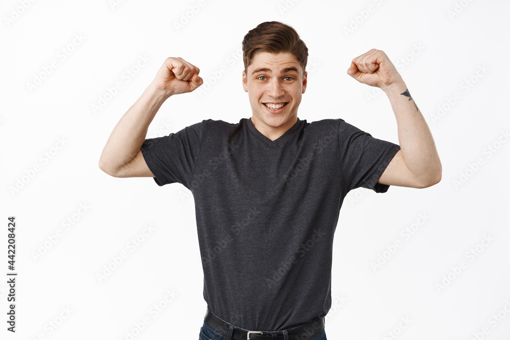 Fototapeta premium Excited young man winning, chanting and celebrating victory, raising hands up in hooray triumph gesture, achieve goal and fist pump, standing happy against white background