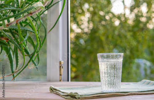 A glass glass with mineral water with gas bubbles stands on a wooden window sill with the window open to the street. Topic thirst quenching, a healthy natural drink