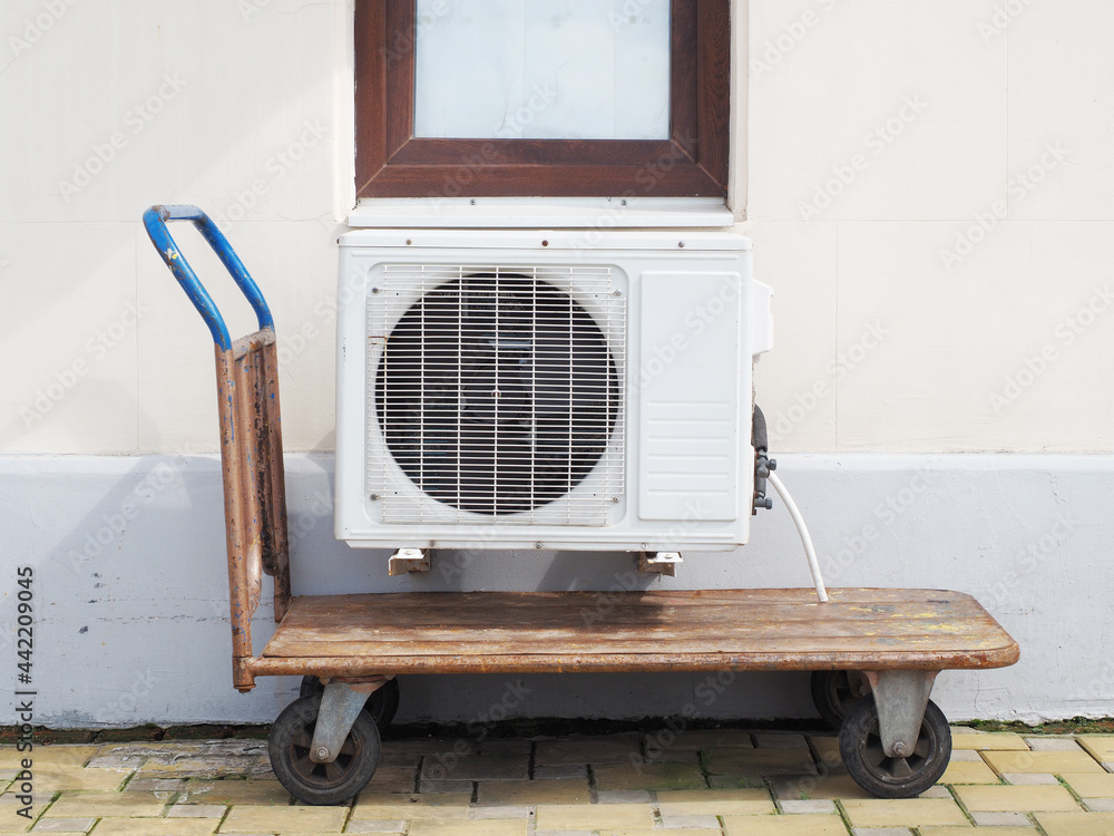 The air conditioner hangs on the wall of the building outside above the pushcart standing on the tile The air conditioner hangs on the wall of the building outside above the pushcart standing on the tile