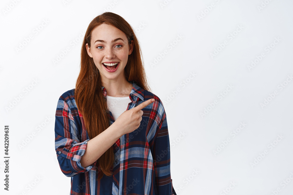 Excited redhead girl with straight hair and white smile, pointing finger right at logo, showing advertisement on empty space, standing against white background Excited redhead girl with straight hair and white smile, pointing finger right at logo, showing advertisement on empty space, standing against white background