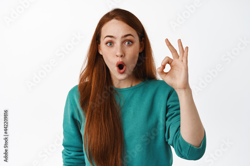Surprised and impressed ginger girl look in awe, gasp and say wow, show okay zero gesture, OK sign, praise and compliment great thing, white background