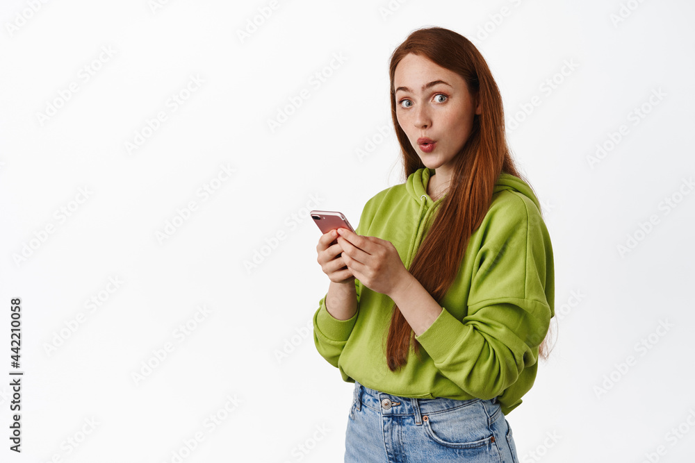 Excited teen redhead girl chatting on mobile phone, holding smartphone and look surprised at camera, standing over white background Excited teen redhead girl chatting on mobile phone, holding smartphone and look surprised at camera, standing over white background