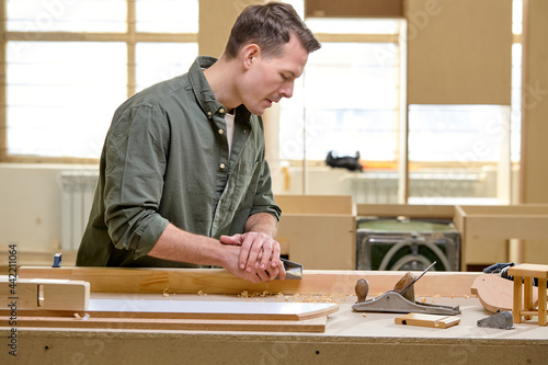 Joiner's works: confident male carpenter scribbles the board with planer, in workshop, dressed casually, side view portrait of handsome hardworking male engaged in woodworking. copy space
