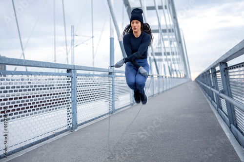 Young fitness woman in winter blue sportswear doing exercise with jump rope on bridge during cold day © pucko_ns