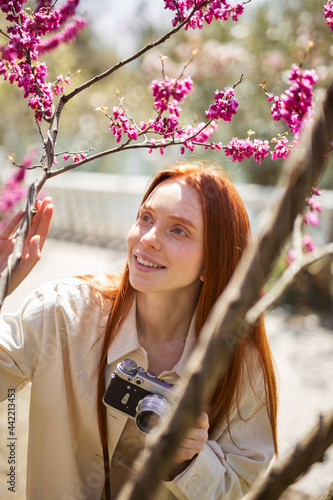 Nature Photographer taking pictures outdoors. Beautiful red-haired caucasian woman between flowering trees with camera, enjoy beauty around in nature. Copy space, people lifestyle concept