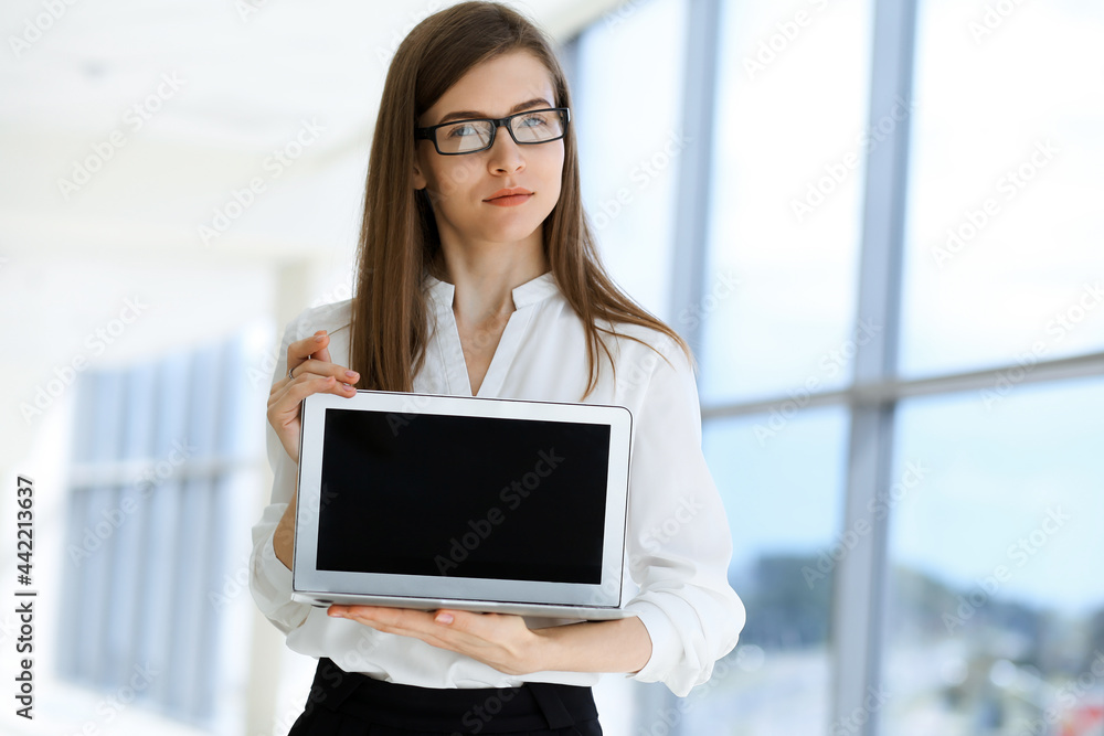 Beautiful female specialist with laptop computer standing in modern office and smiling charmingly. Working on design, data analysis, plan strategy. Business people concept Beautiful female specialist with laptop computer standing in modern office and smiling charmingly. Working on design, data analysis, plan strategy. Business people concept