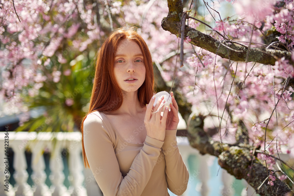 Woman enjoying sunny day in botanical garden, using cream, beauty skin care products. Sensual redhead woman posing under blooming blossom tree, holding cream bottle in hands Woman enjoying sunny day in botanical garden, using cream, beauty skin care products. Sensual redhead woman posing under blooming blossom tree, holding cream bottle in hands