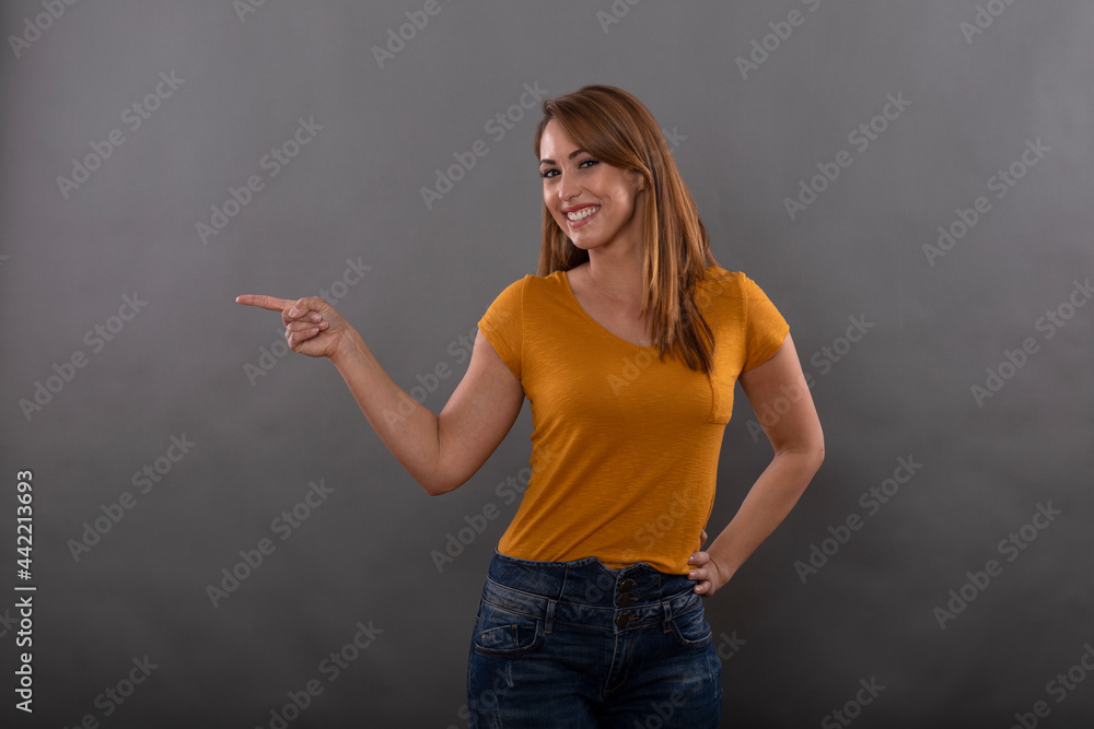 Cheerful brunette model dressed in casual clothes smiles and points sideways with finger isolated on gray background Cheerful brunette model dressed in casual clothes smiles and points sideways with finger isolated on gray background