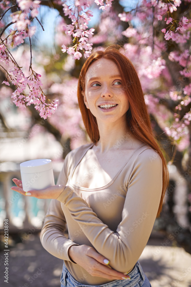 Happy smiling beautiful female with natural makeup posing with cream jar, near blooming flowers. Close up outdoor portrait. Spring, beauty, skin care concept. Copy space Happy smiling beautiful female with natural makeup posing with cream jar, near blooming flowers. Close up outdoor portrait. Spring, beauty, skin care concept. Copy space