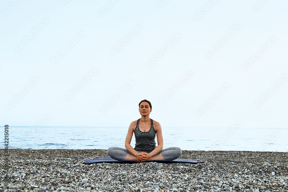 Fototapeta premium Slim Woman doing Padmasana or lotus pose on the beach with at the morning. Strengthening muscles of legs and coordination of body. Practicing yoga outdoors.