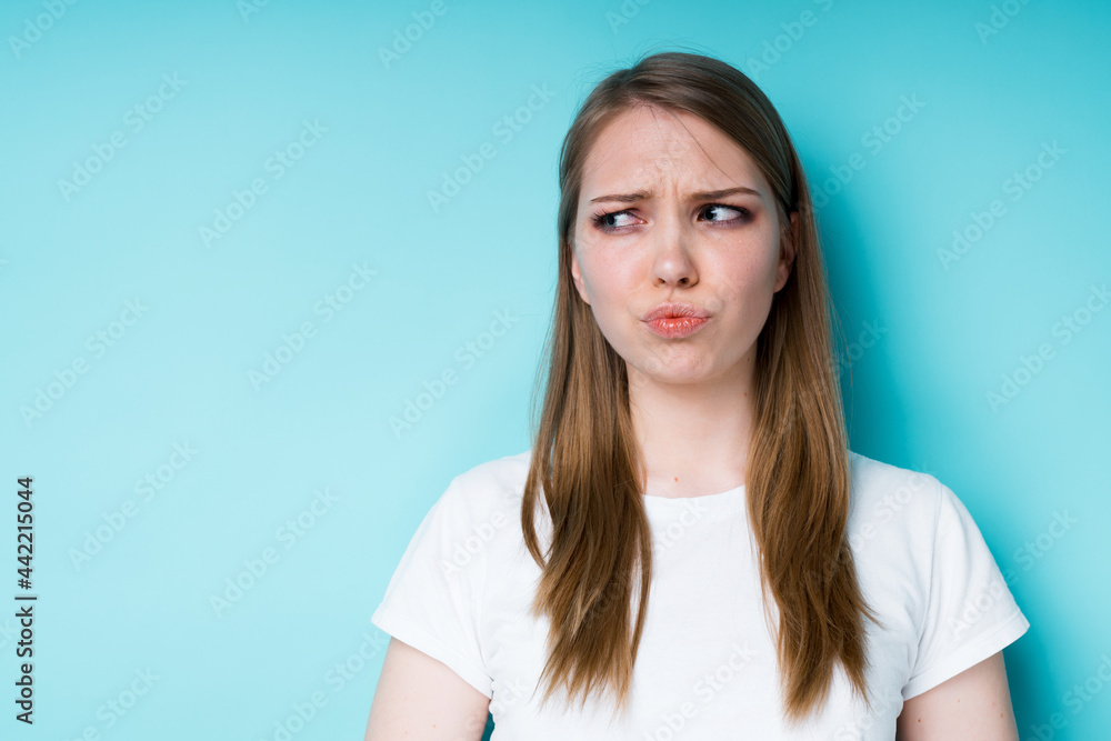 A beautiful girl in a white T-shirt pouts her lips offendedly and looks to the side while standing on a blue background A beautiful girl in a white T-shirt pouts her lips offendedly and looks to the side while standing on a blue background
