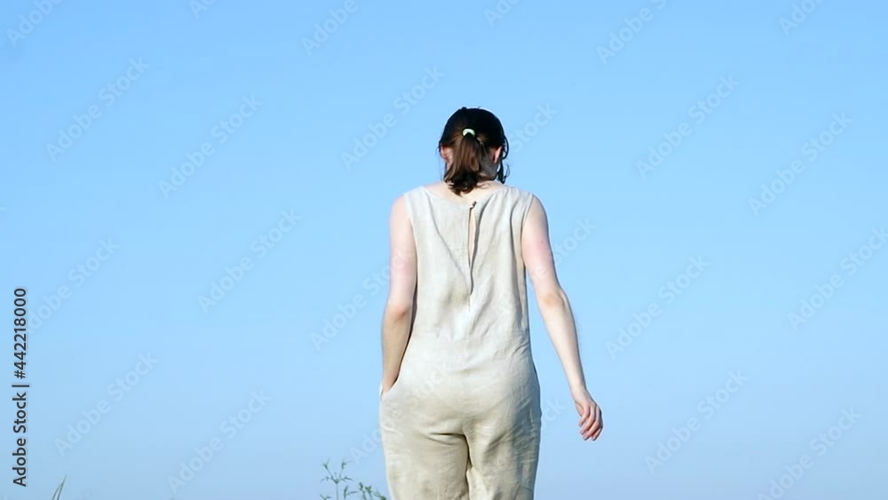 Woman in handmade natural linen jumpsuit wearing sunglasses running away from the frame, onto a hilltop, against blue sky Woman in handmade natural linen jumpsuit wearing sunglasses running away from the frame, onto a hilltop, against blue sky