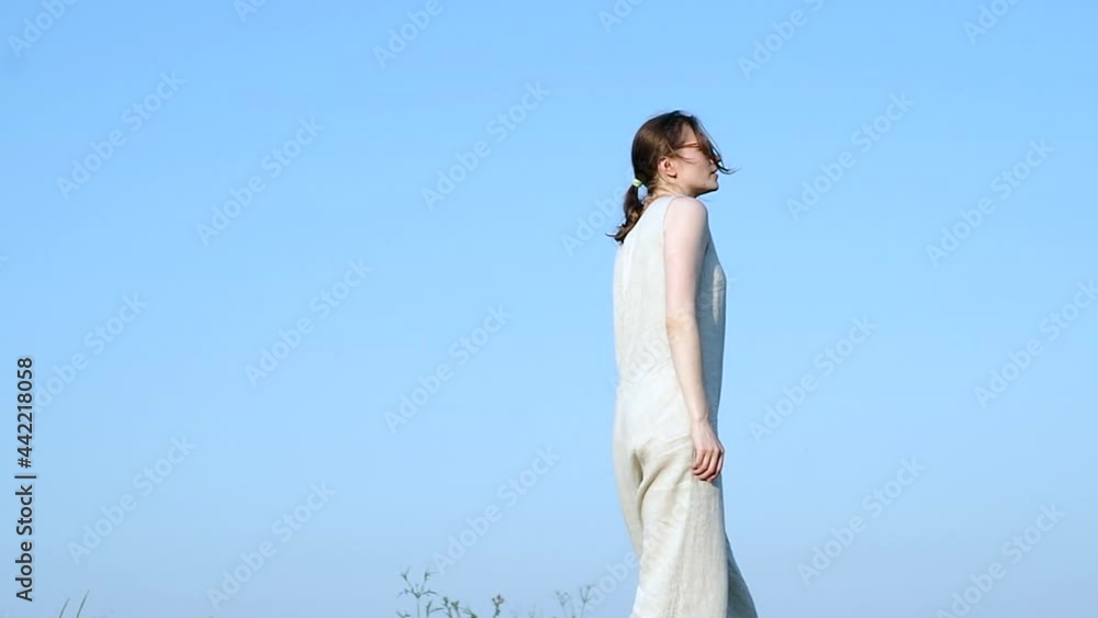 Woman in handmade natural linen jumpsuit wearing sunglasses walking on a hill, against blue sky, arms to the side, portraying freedom, ease, carelessness. Woman in handmade natural linen jumpsuit wearing sunglasses walking on a hill, against blue sky, arms to the side, portraying freedom, ease, carelessness.