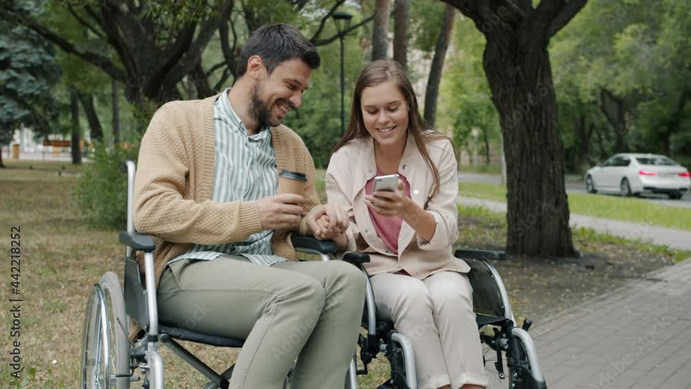 Slow motion of disabled young people couple talking and using smartphone sitting in wheelchairs in park enjoying leisure time outdoors. Youth and disability concept. Slow motion of disabled young people couple talking and using smartphone sitting in wheelchairs in park enjoying leisure time outdoors. Youth and disability concept.