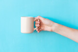 Human hand holding big ceramic beige mug above light blue background with copy space. Mug with empty clean surface.
