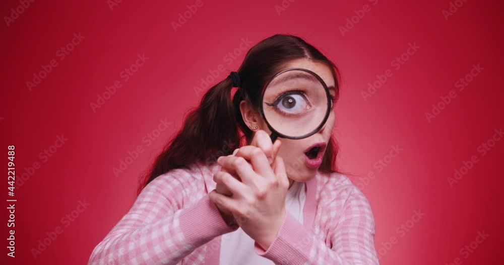 Young funny attractive caucasian girl with long braids holding a magnifying glass in front of camera and makes faces and grimaces on red backgorund. Human curiosity search concept Young funny attractive caucasian girl with long braids holding a magnifying glass in front of camera and makes faces and grimaces on red backgorund. Human curiosity search concept