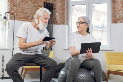 Senior couple in sport clothes resting at living room with digital tablet and smartphone in hands. Aged man and woman drinking water and surfing internet after training.