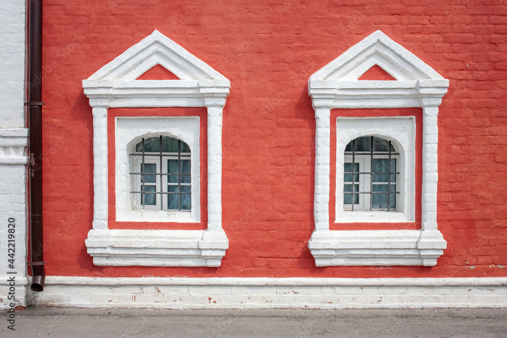 Fototapeta premium Two windows on the red wall of the house. Decor and framing of window openings on the facade. Window openings with a semicircular arched finish, included in a rectangular frame.