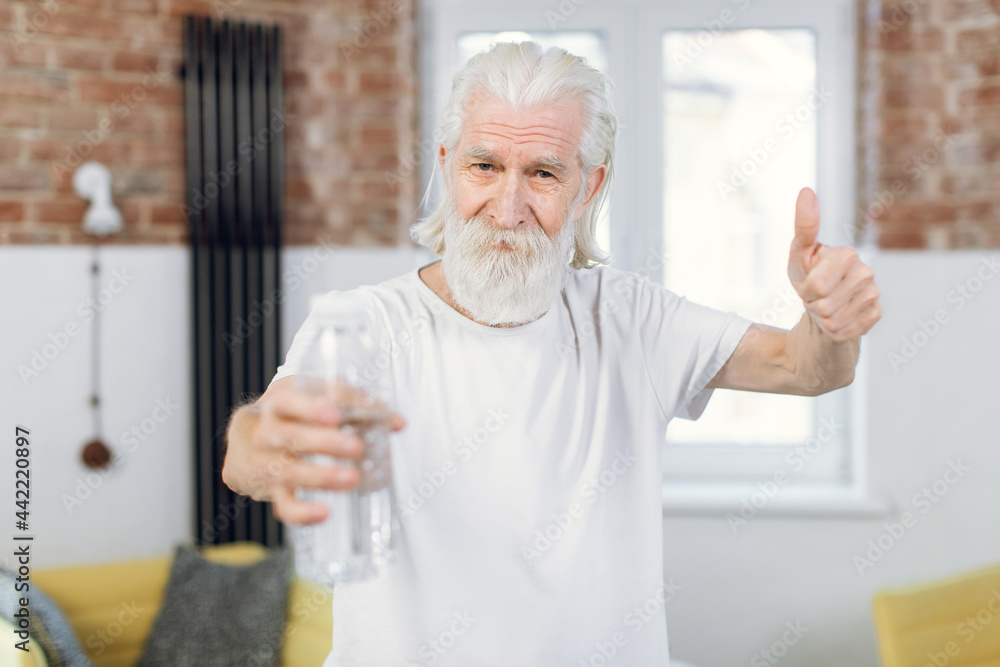 Bearded senior man showing thumb up while holding bottle of fresh water. Male in activewear renewing water balance after workout at home. Bearded senior man showing thumb up while holding bottle of fresh water. Male in activewear renewing water balance after workout at home.