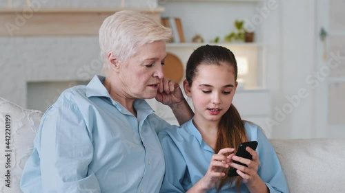 Little cute granddaughter holds smartphone in hands teaches old grandmother to use mobile application, play games and shows photos. Elderly aged babysitter helps girl with education video on cellphone photo