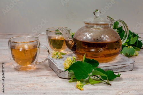 A glass teapot and two cups of tea with a linden tree on a wooden table against a background of linden leaves and flowers. Healing tea.