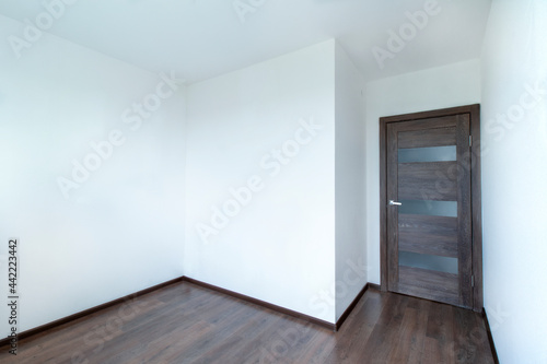 A room with white walls with right angles, a wooden brown door and a dark laminate floor. Without a window. The four corners of the room are visible.