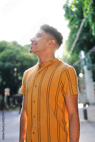 Young gay latino man wearing a yellow striped shirt with clipping light and trees in the background
