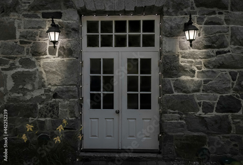 White metal doors and transom window on a grey natural stone building, illuminated by two lantern style exterior lights, nobody
