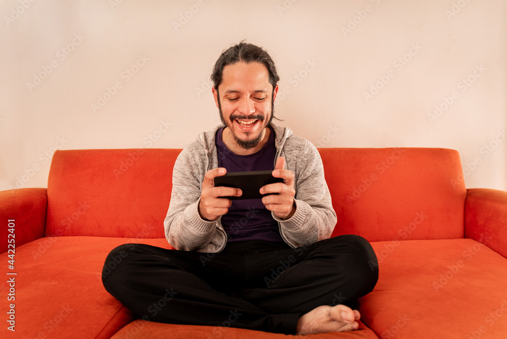 Young man playing gaming on the smart phone sitting on an orange sofa couch at home in the living room Young man playing gaming on the smart phone sitting on an orange sofa couch at home in the living room