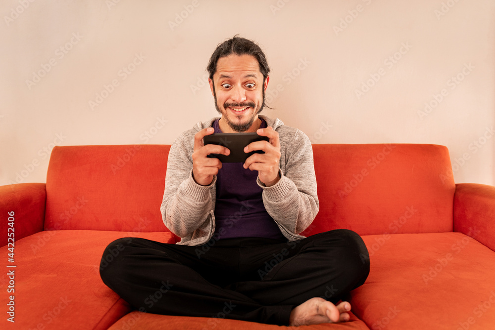 Young man playing gaming on the smart phone sitting on an orange sofa couch at home in the living room