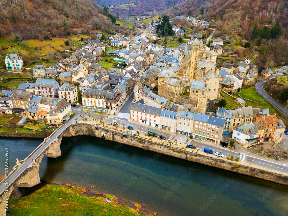 Obraz premium Aerial view of ancient castle of Chateau Estaing and medieval arched bridge across river Lot on background of Estaing cityscape in cloudy winter day, Aveyron, France..