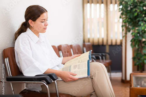 Focused young girl sits in the lobby of a private medical clinic and attentively reads a magazine