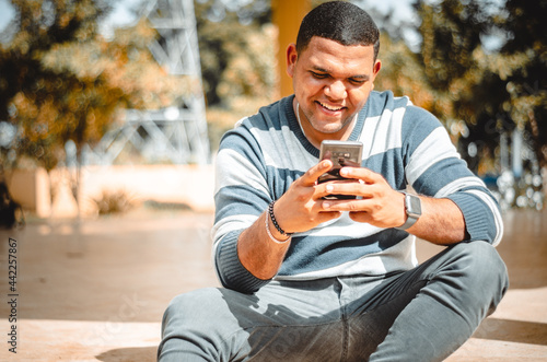 latin dominican man texting with his phone while smiling, sitting in the park photo