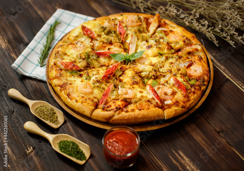 Selective focus and close up on homemade seafood pizza with shrimp, pineapples, bell pepper, crab sticks on wooden tray and table decorated with tomato sauce or ketchup, dried plants and oregano.