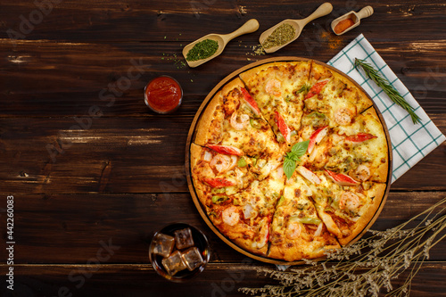 Close up on homemade seafood pizza with shrimp, bell pepper, crab sticks on wooden table with cold beverage, napkin, ingredient of tomato sauce or ketchup, dried plants, paprika chili powder, oregano.