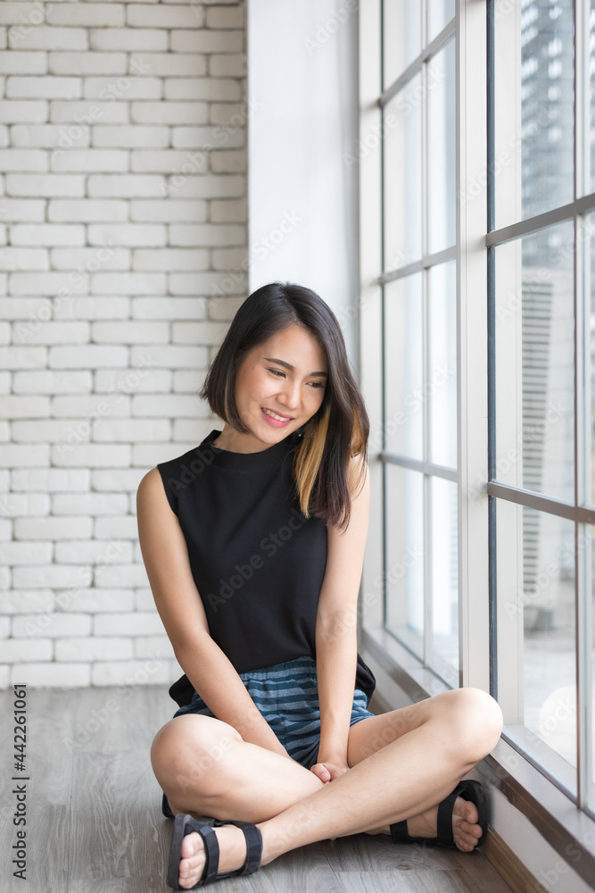 Horizontal portrait shot of a cute smiling Asian woman with a straight hairstyle in black clothes sitting near a window in the studio with a Christmas tree foreground Horizontal portrait shot of a cute smiling Asian woman with a straight hairstyle in black clothes sitting near a window in the studio with a Christmas tree foreground