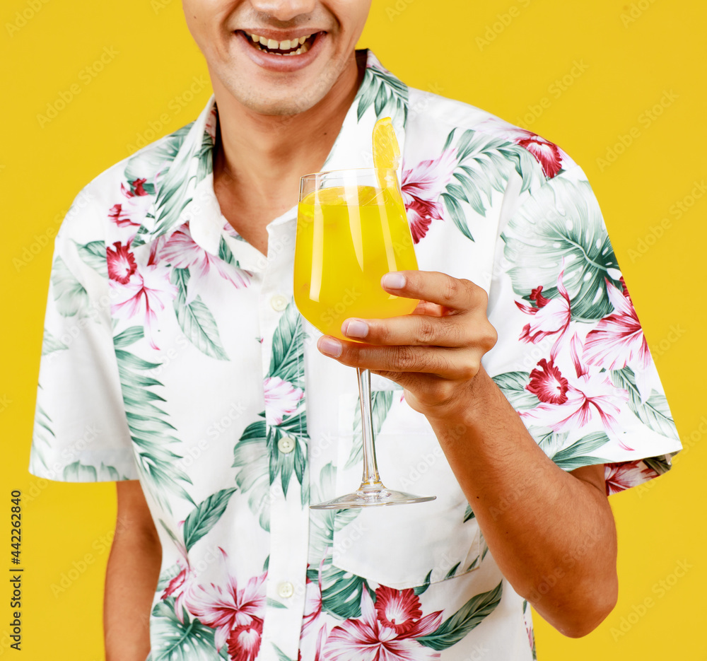 Young attractive Asian man in white Hawaiian shirt holding a glass of orange juice against yellow background. Concept for beach vacation holiday Young attractive Asian man in white Hawaiian shirt holding a glass of orange juice against yellow background. Concept for beach vacation holiday